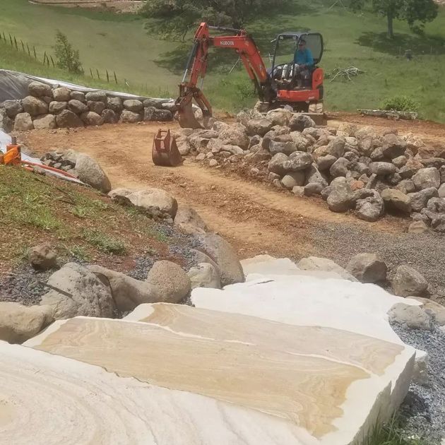 A Red Excavator Is Moving Rocks On A Dirt Road — Full Circle Landscaping in Uki, NSW