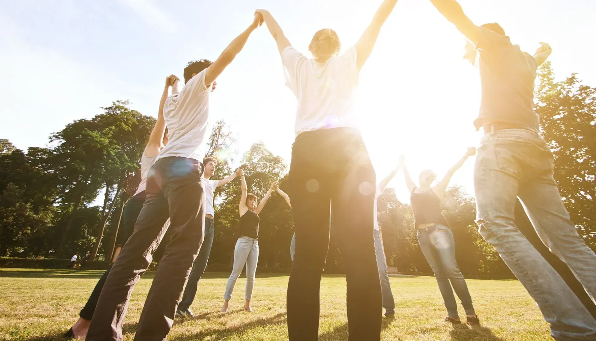 People holding hands in a circle outdoors, arms raised. Sunlight shines behind them in a park setting.