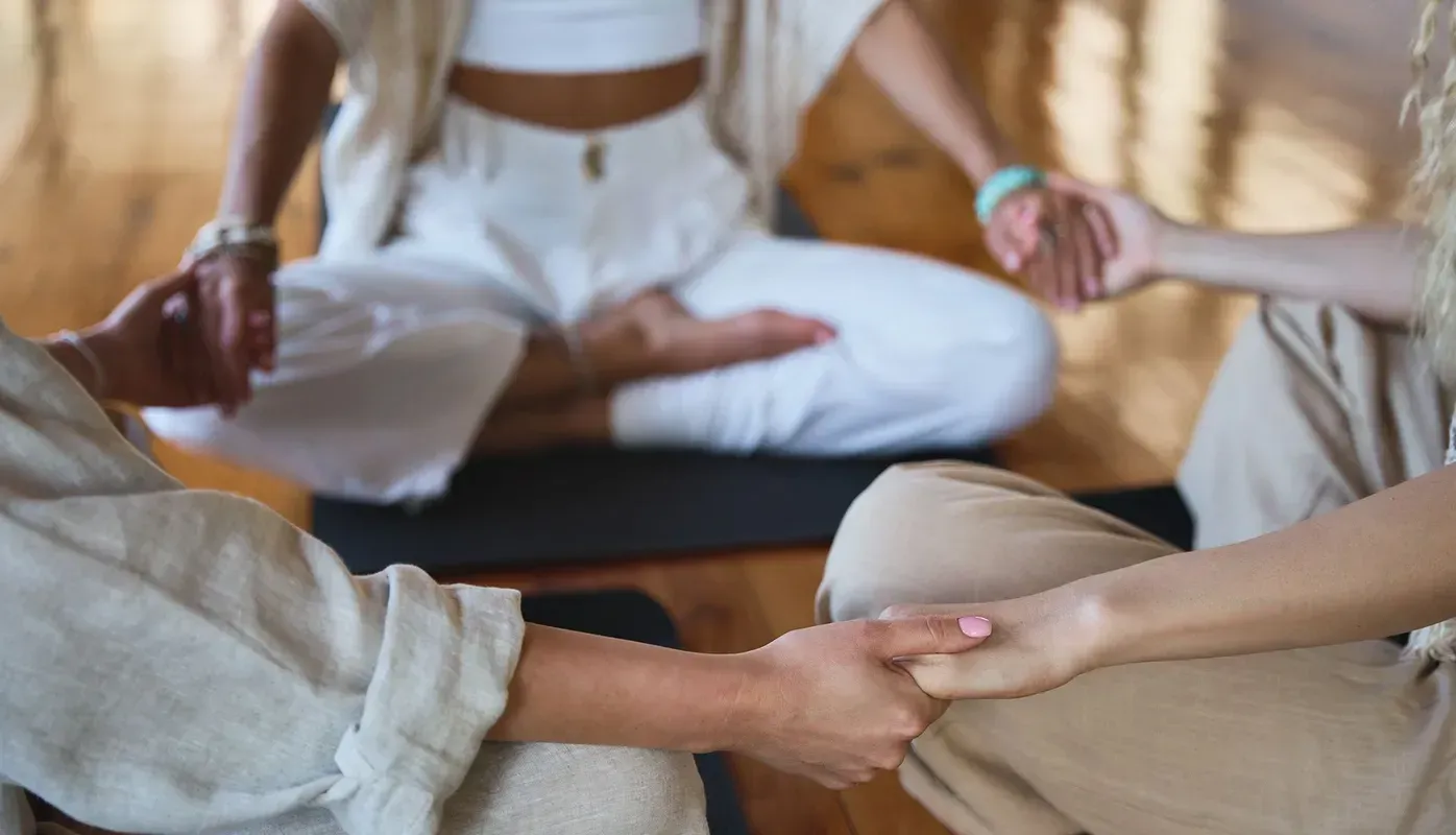 People sitting in a circle, holding hands. One person is in a lotus position, indoors, neutral tones.