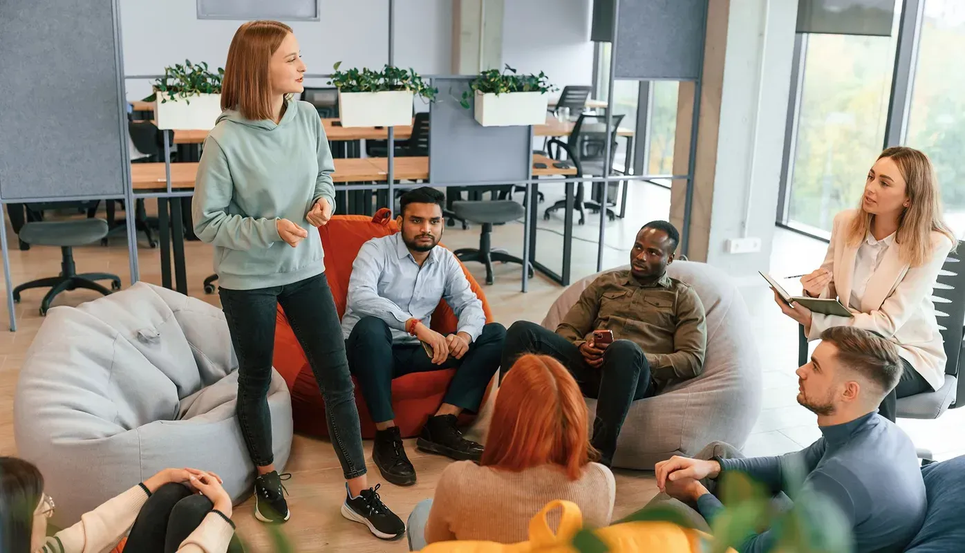 Group in a modern office setting, listening to a presentation. Woman speaks, others seated on beanbags and chairs.