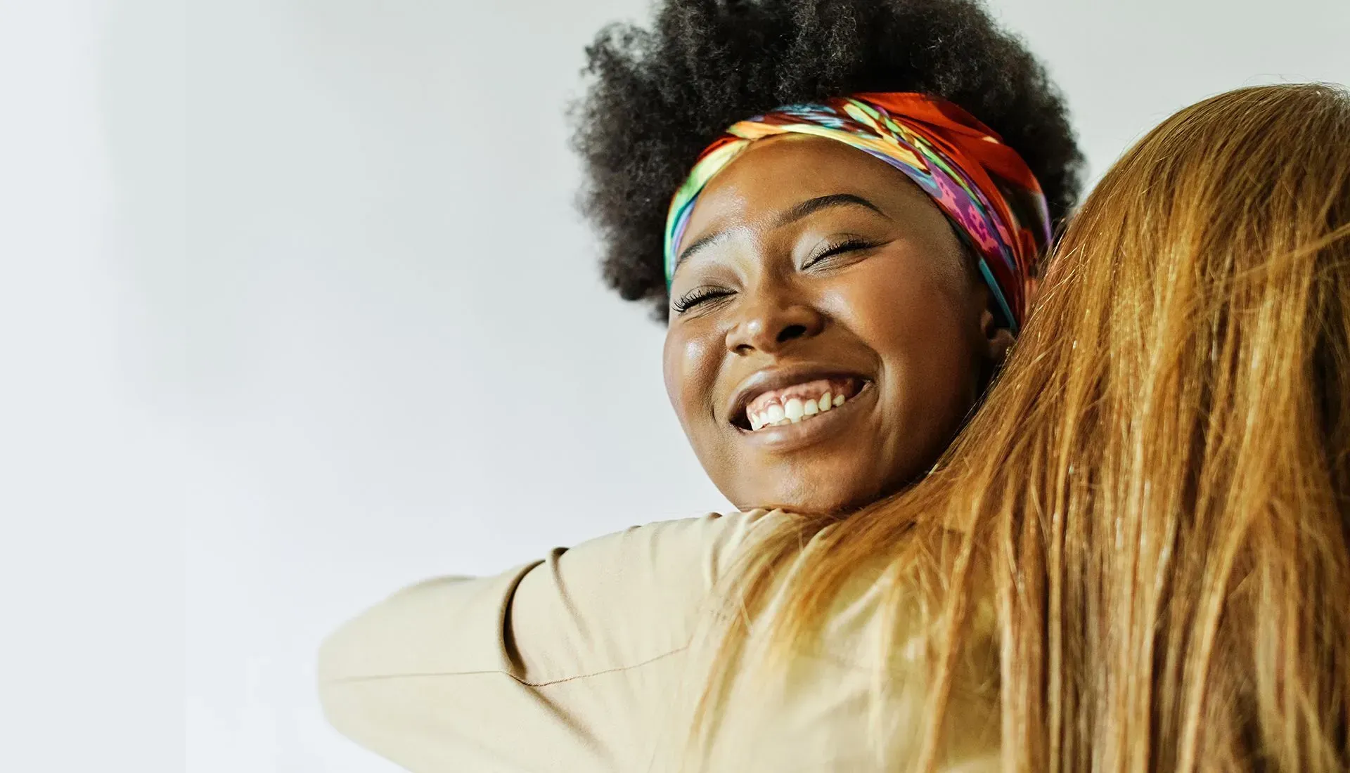 Smiling woman with colorful headband hugs another person, both against a white background.