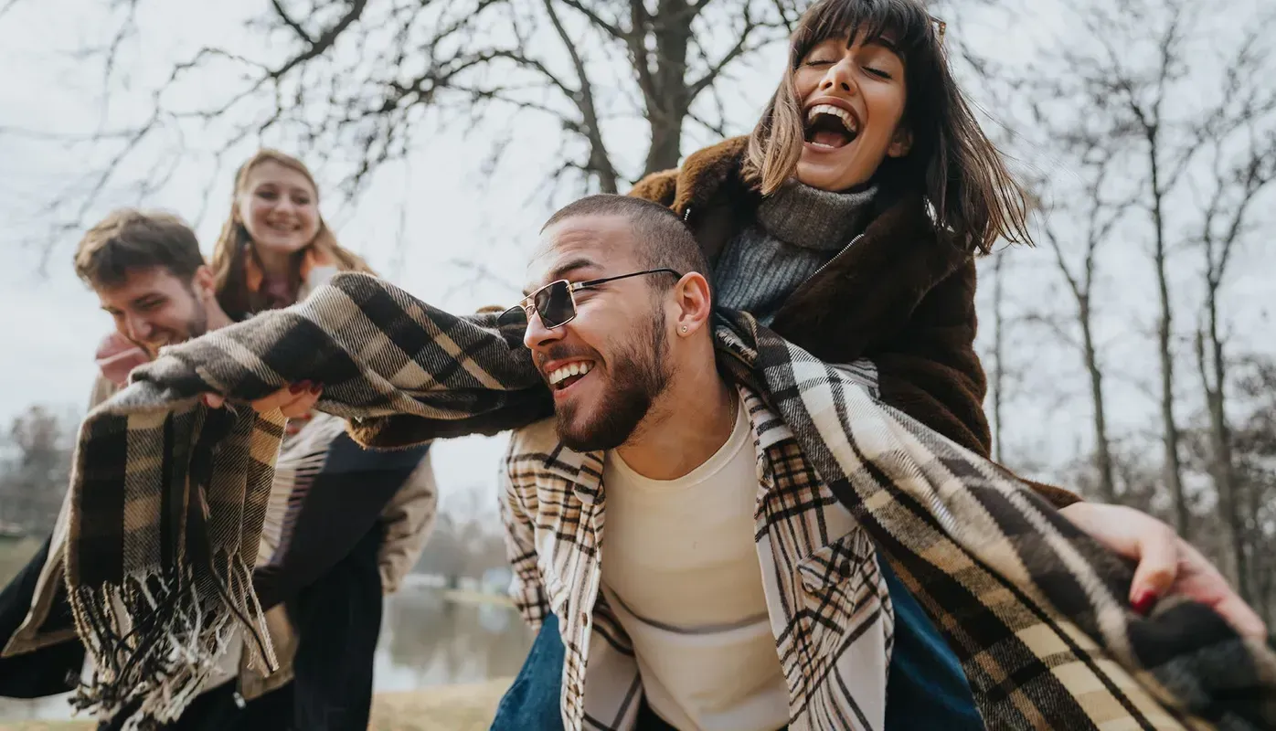 Four people laughing, playfully piggybacking in a park, enjoying an outdoor outing.
