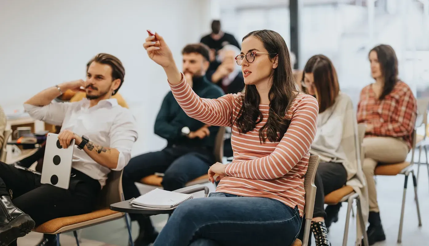 Woman in a striped shirt raising hand in a classroom, pointing with a pen. Others sit and listen.