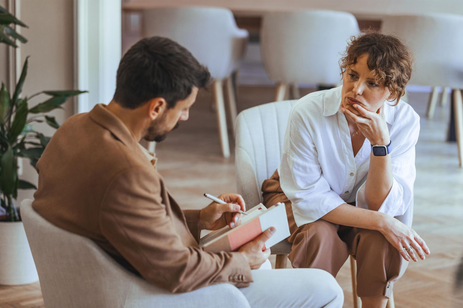 Therapist taking notes while a woman with hand on chin looks concerned in an office.