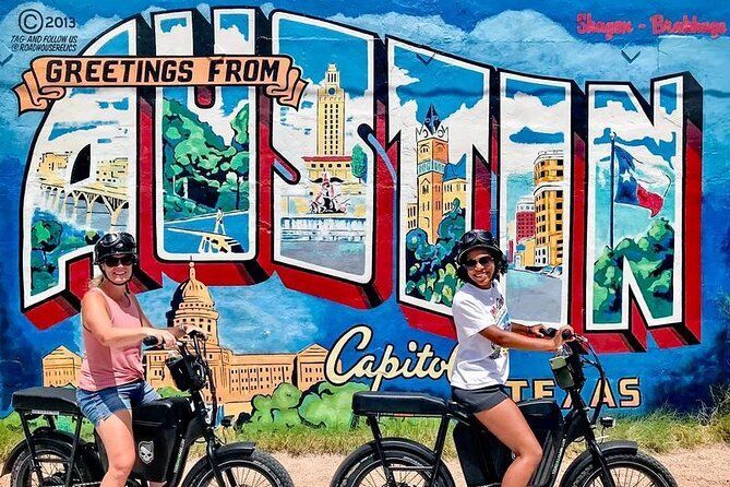 Two women are riding bicycles in front of a mural that says greetings from austin.