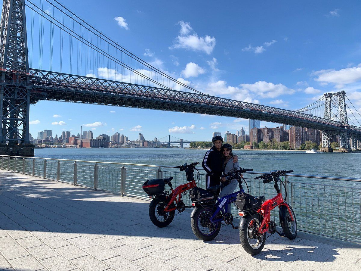 A couple standing next to two bicycles in front of a bridge over a body of water in NY