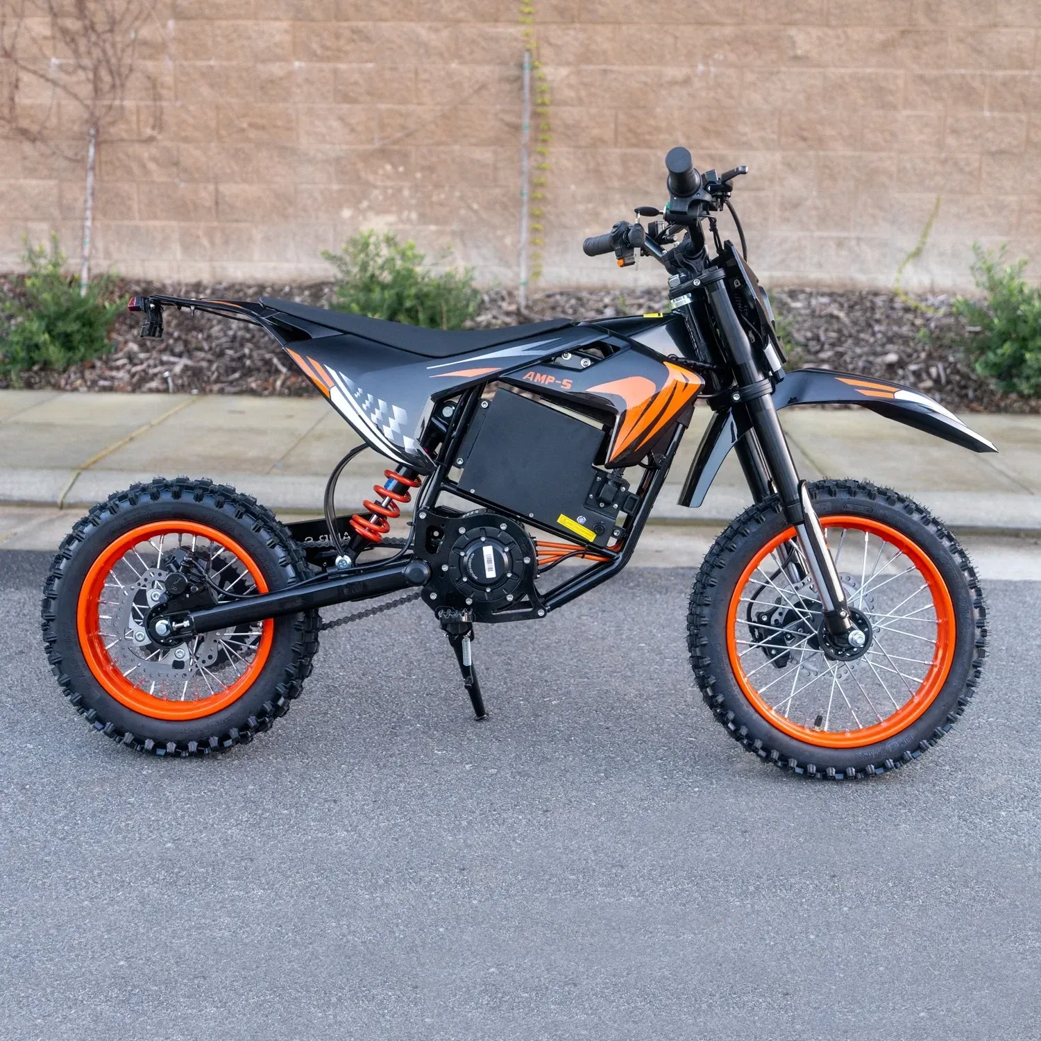 Black and orange electric dirt bike with knobby tires parked on asphalt against a concrete wall.