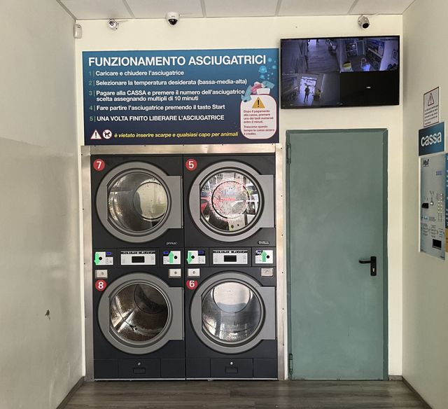 Lavanderia self-service con quattro lavatrici a carica frontale, cartello informativo, monitor CCTV e porta verde.