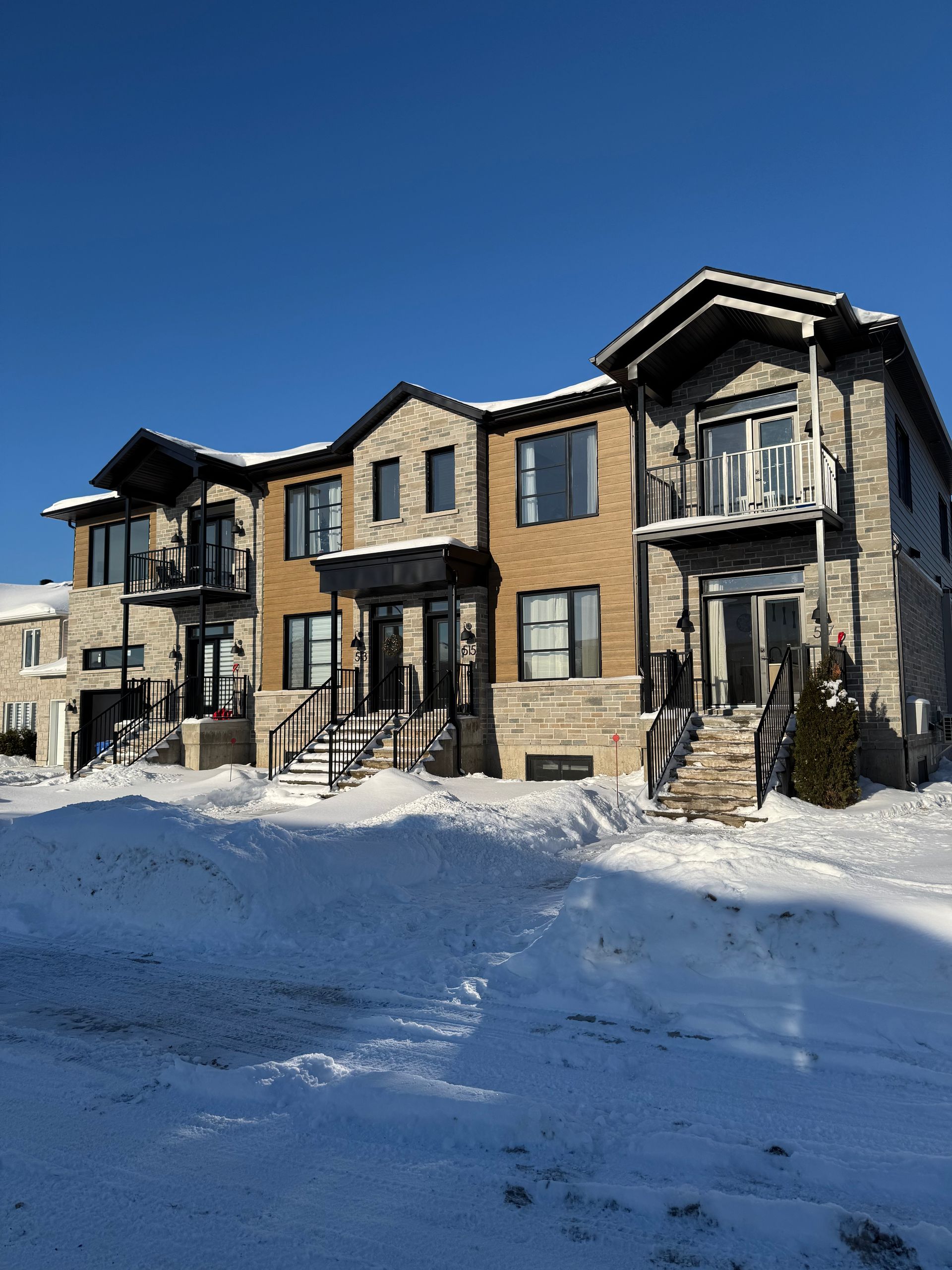 Maisons de ville en hiver, avec sol enneigé, façades en pierre et en brique, portes sombres et balcons sous un ciel bleu limpide.