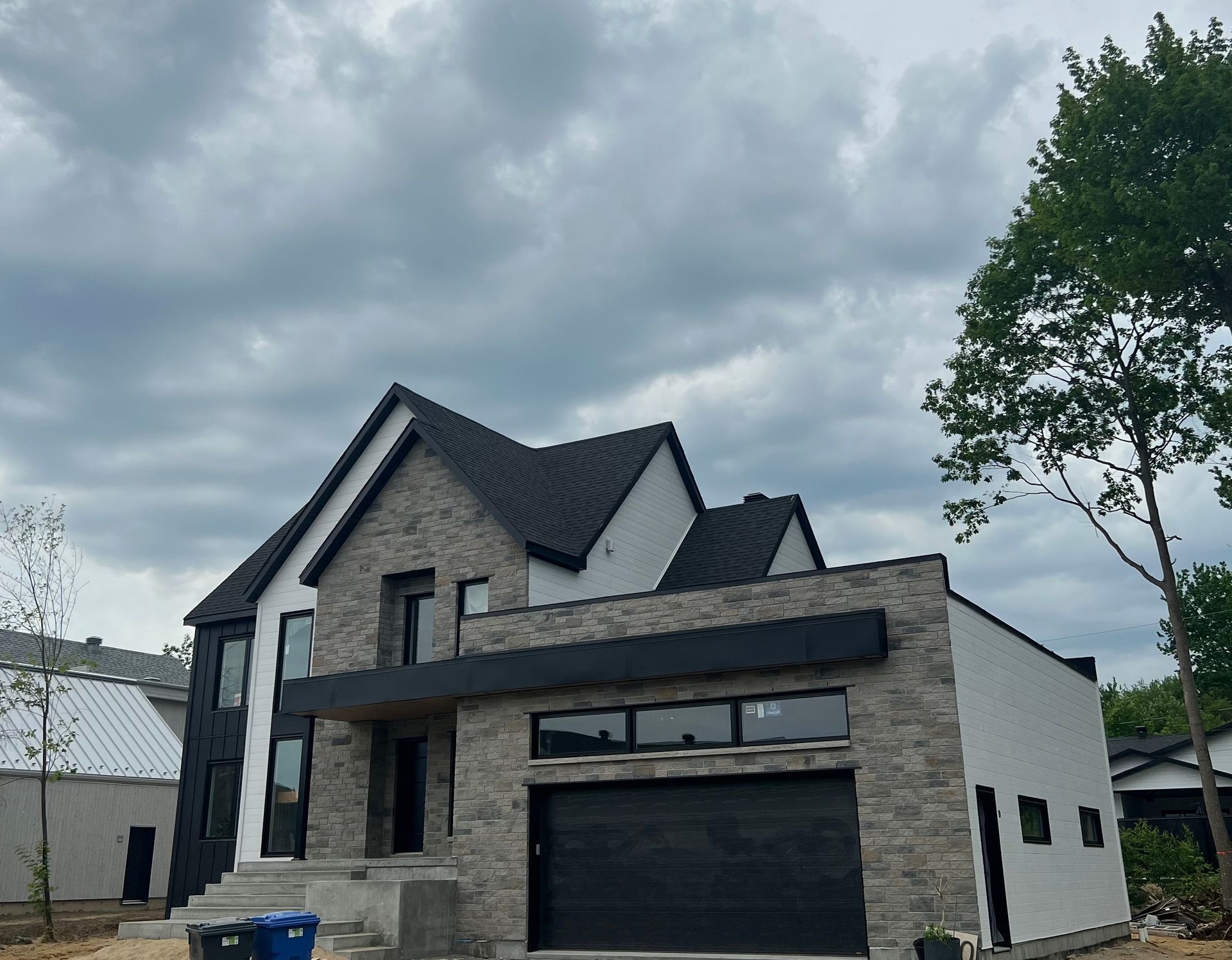 Maison moderne à deux étages avec façade en pierre et bardage noir, porte de garage noire, sous un ciel nuageux.