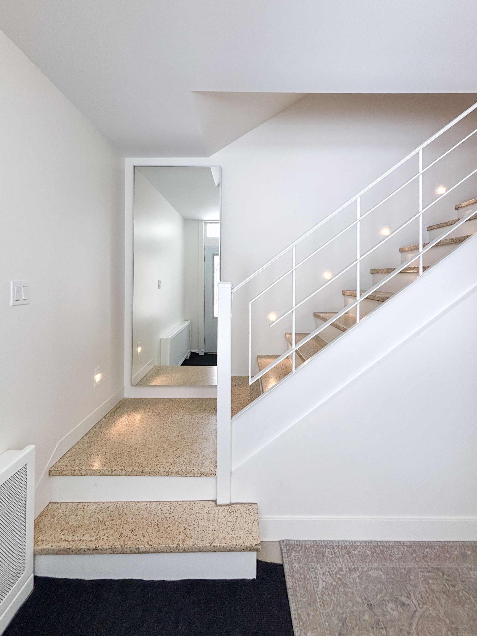Escalier avec rampe blanche, marches à surface mouchetée, grand miroir à gauche reflétant un couloir.