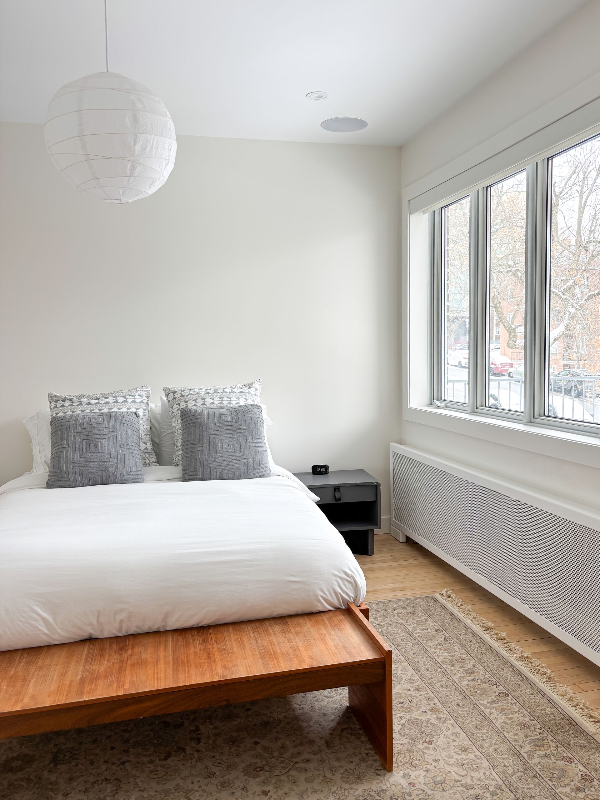 Chambre aux murs blancs, lit avec draps blancs et oreillers gris. Un banc en bois et un tapis devant.