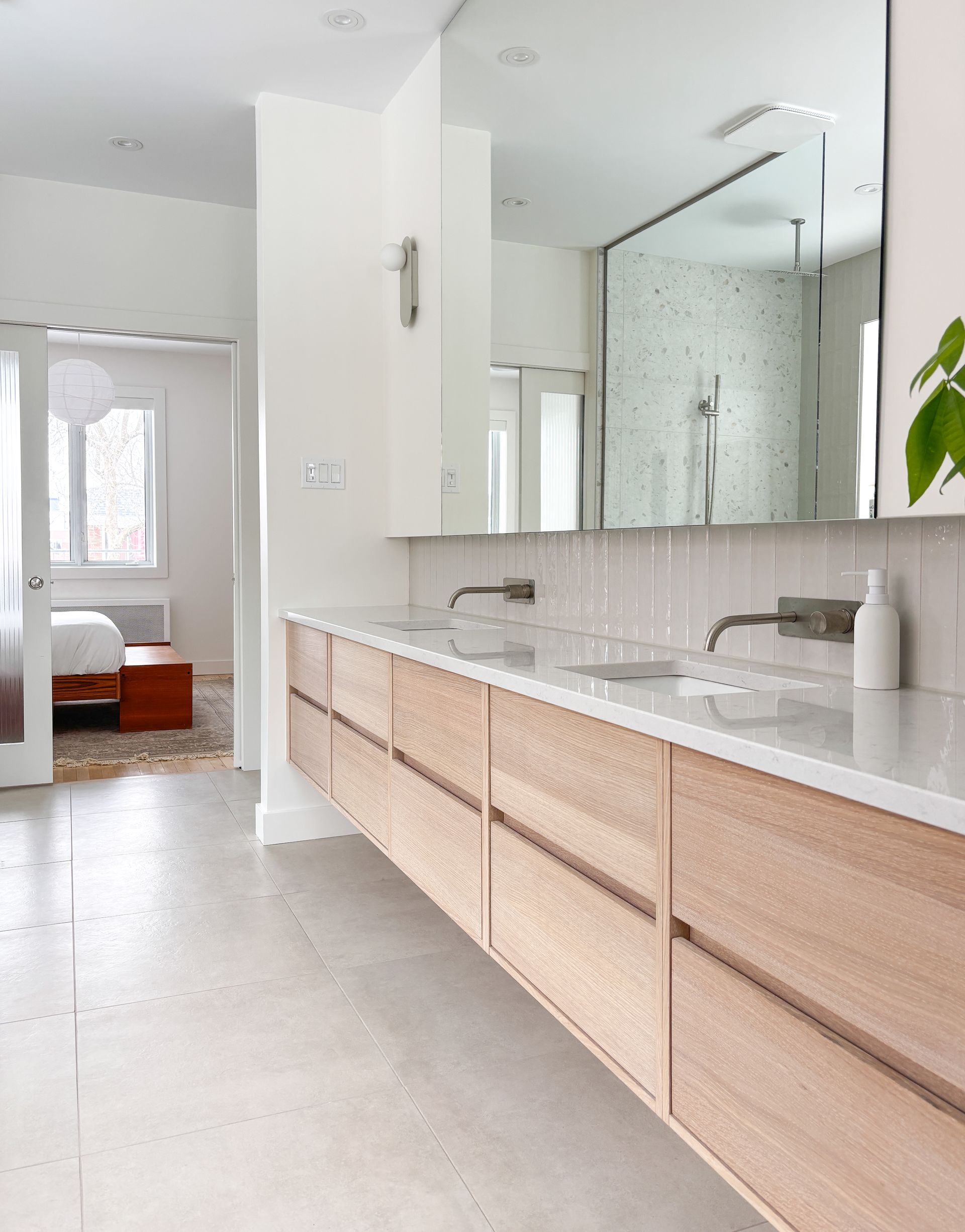 Salle de bain spacieuse et moderne avec vanité flottante en bois clair, grand miroir et porte ouverte sur une chambre.