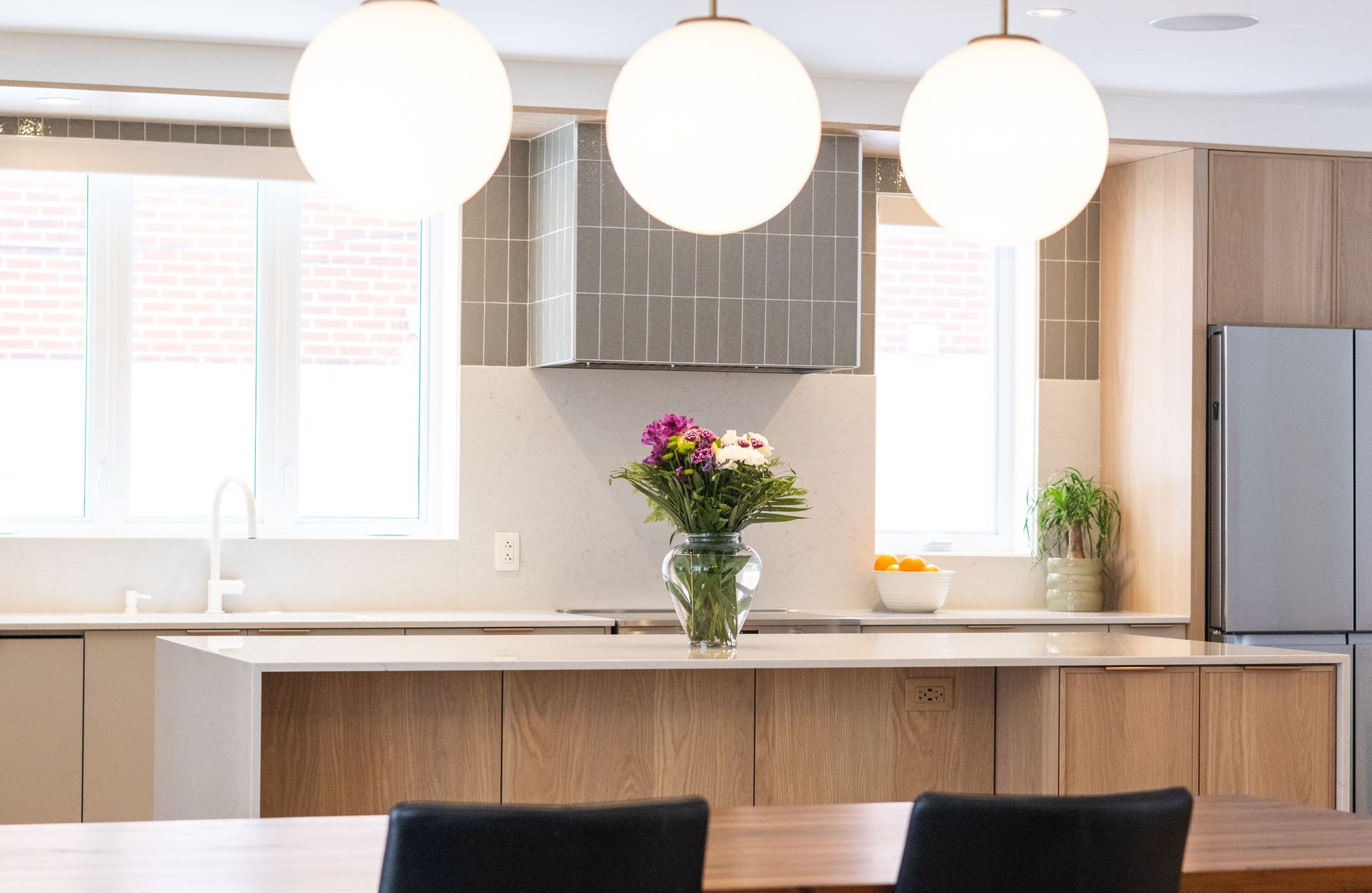 Cuisine moderne avec îlot, luminaires suspendus, armoires en bois, réfrigérateur en acier inoxydable et fleurs.