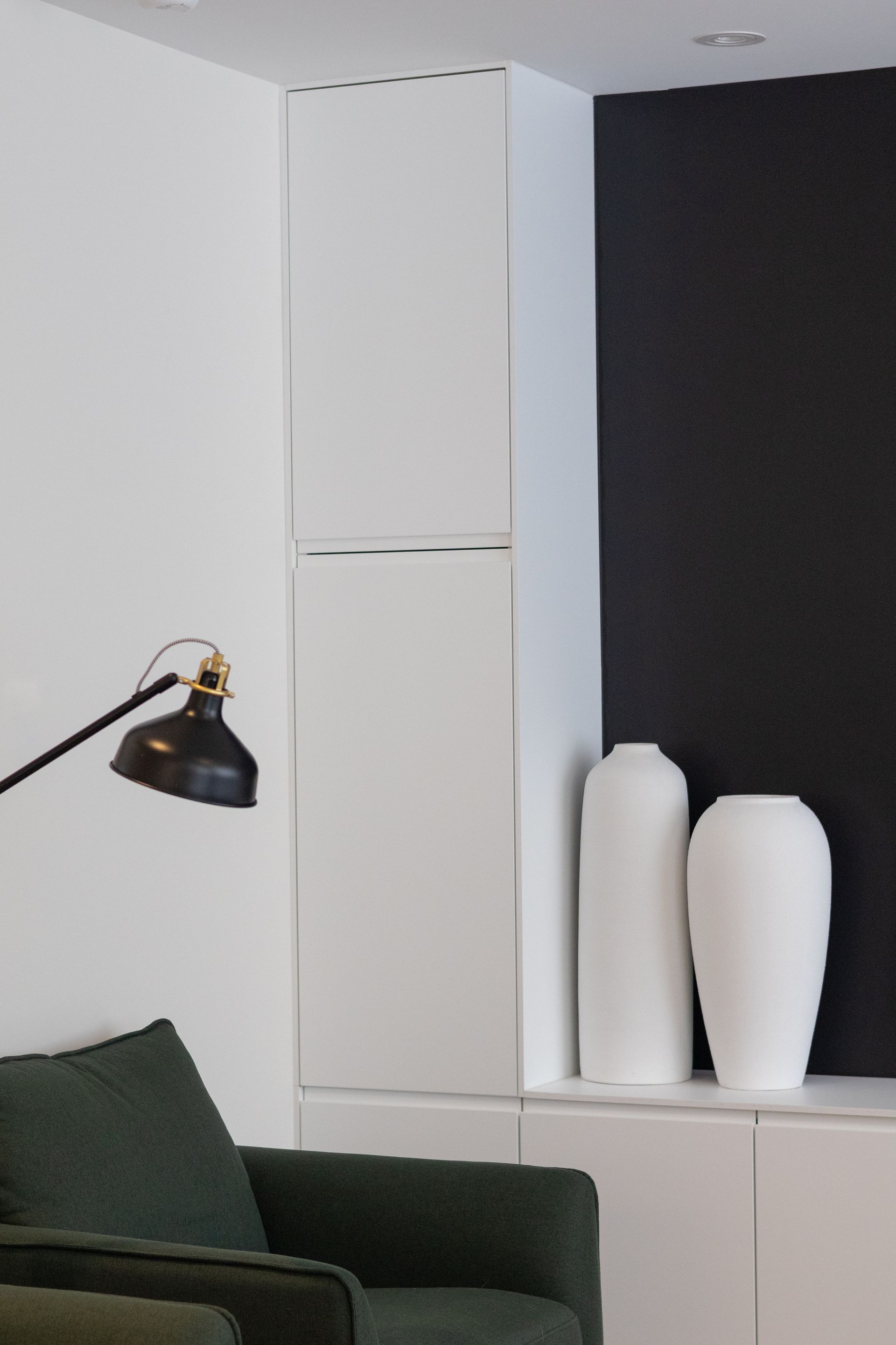 Vases blancs sur une armoire blanche, devant un mur noir. Une lampe noire et un fauteuil vert sont au premier plan.