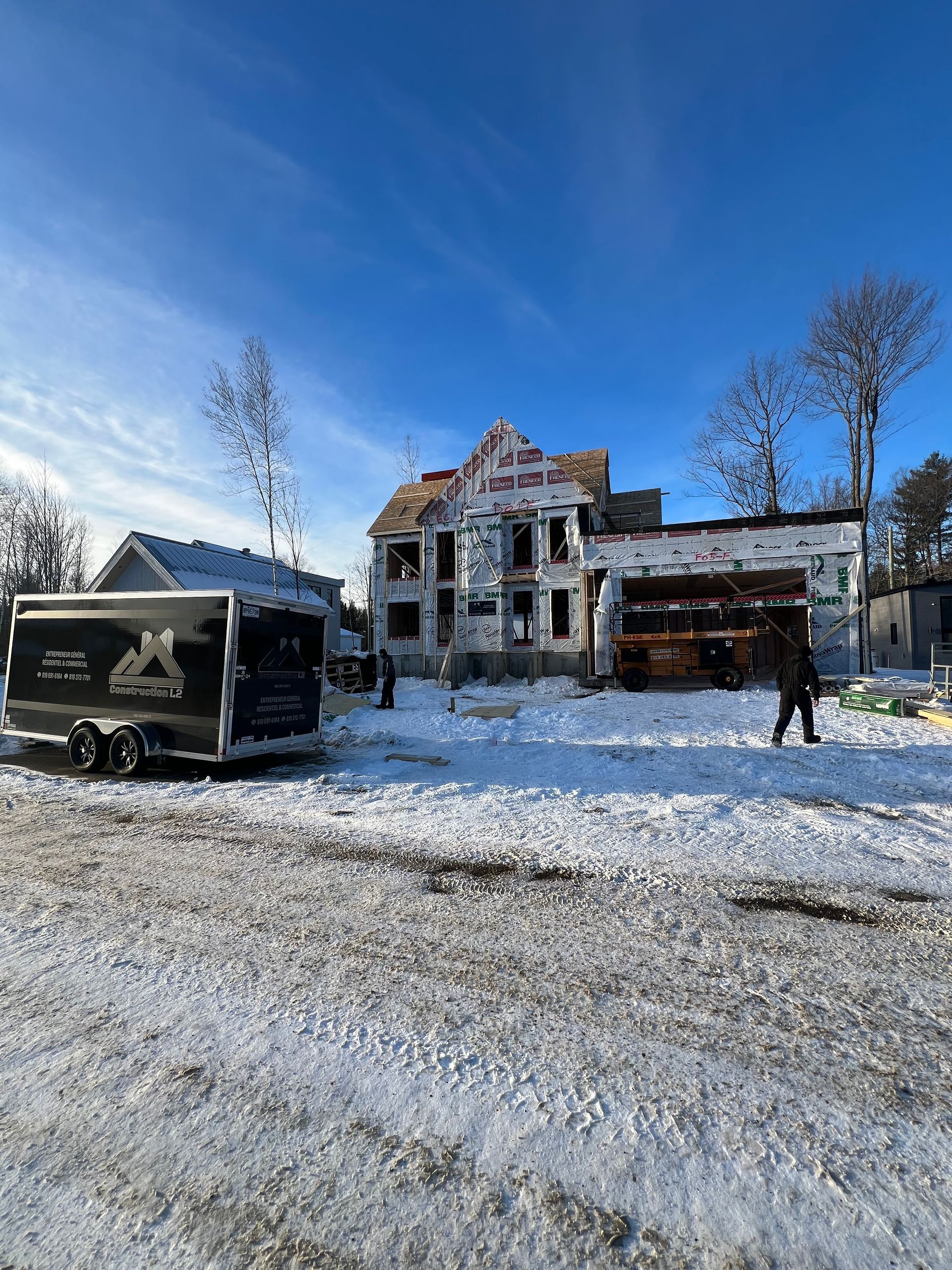 Maison en construction dans un champ enneigé ; caravane et personnes visibles. Ciel bleu.