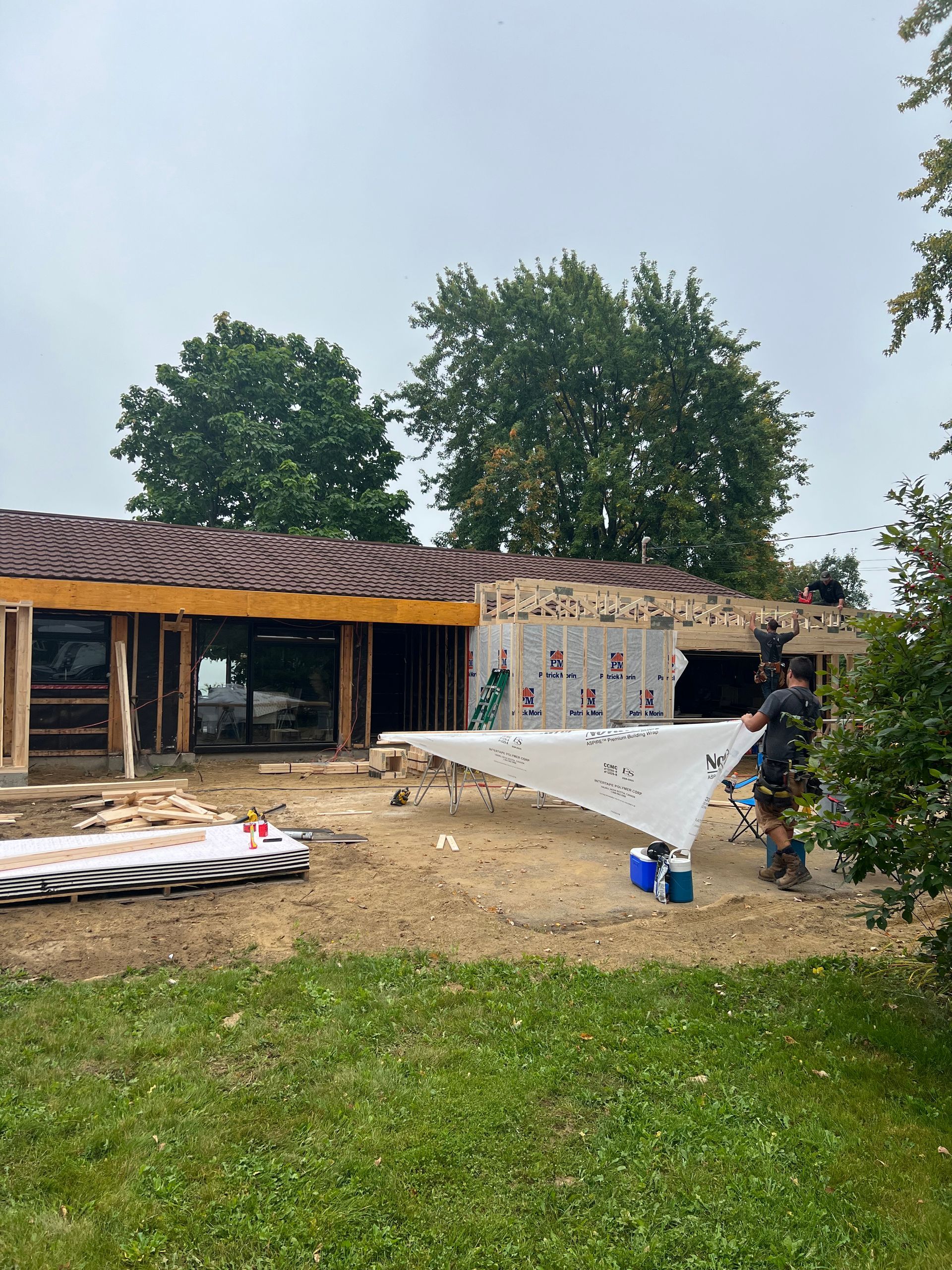 Chantier de construction : des ouvriers installent une bâche blanche sur un bâtiment partiellement construit, dont la charpente en bois est apparente et la toiture brune.