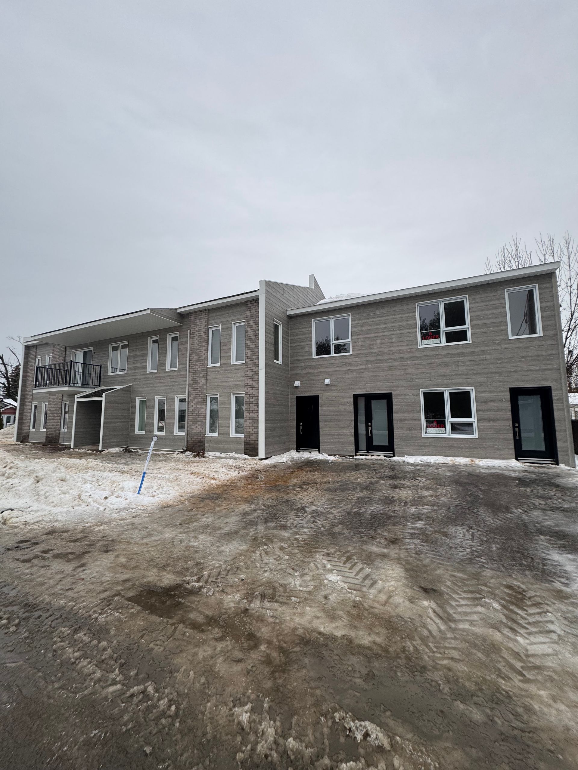 Immeuble d'appartements gris de deux étages avec façade en pierre, neige au sol devant, ciel nuageux.