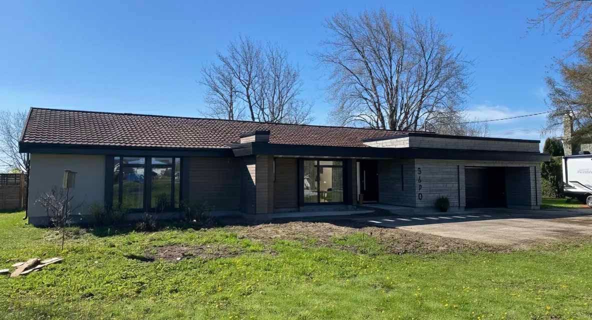 Une maison de plain-pied, discrète, avec un toit plat, de grandes fenêtres et une porte de garage, située dans un jardin gazonné sous un ciel bleu.