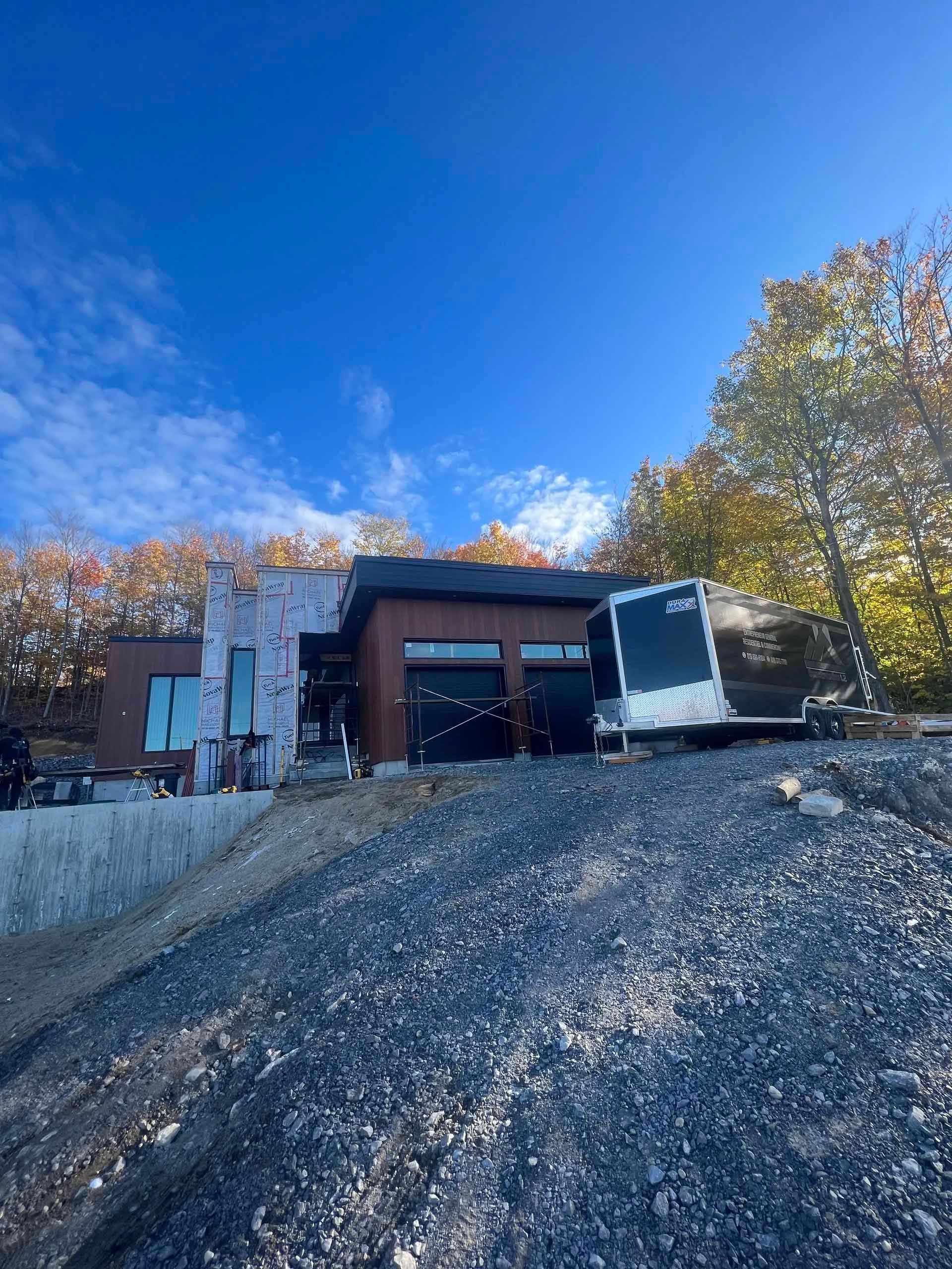 Maison moderne en construction sur une colline de gravier, avec une caravane. Ciel bleu, feuillage d'automne.
