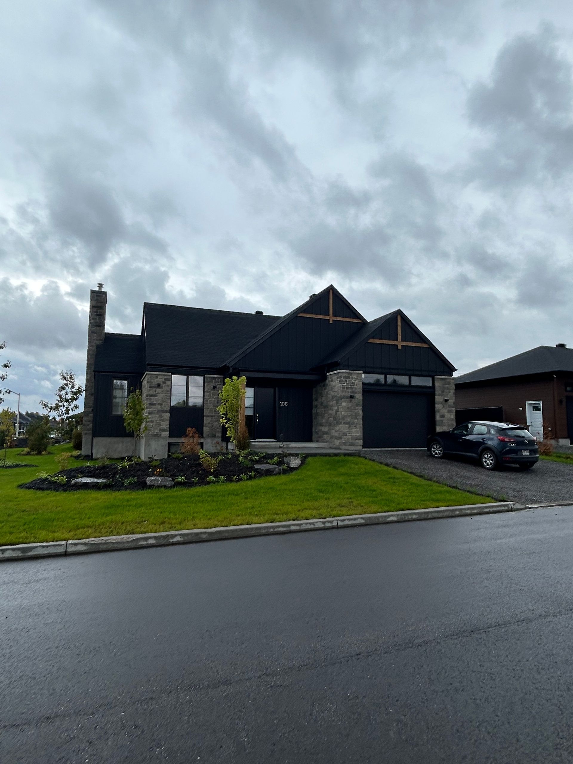 Maison moderne à l'extérieur noir, façade en pierre et toit sombre sous un ciel nuageux.
