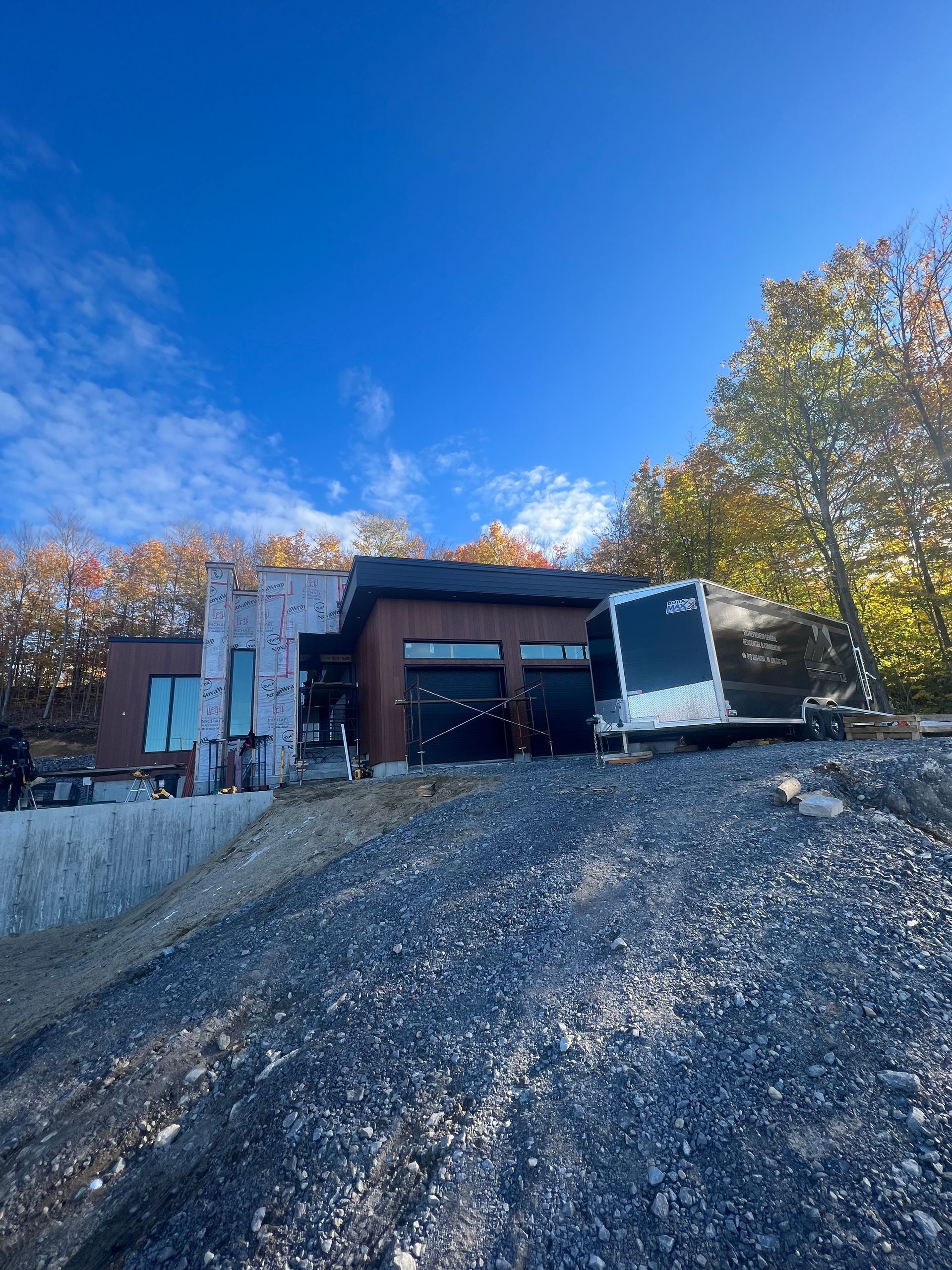 Maison moderne en construction sur une colline de gravier, avec une caravane. Ciel bleu, feuillage d'automne en arrière-plan.