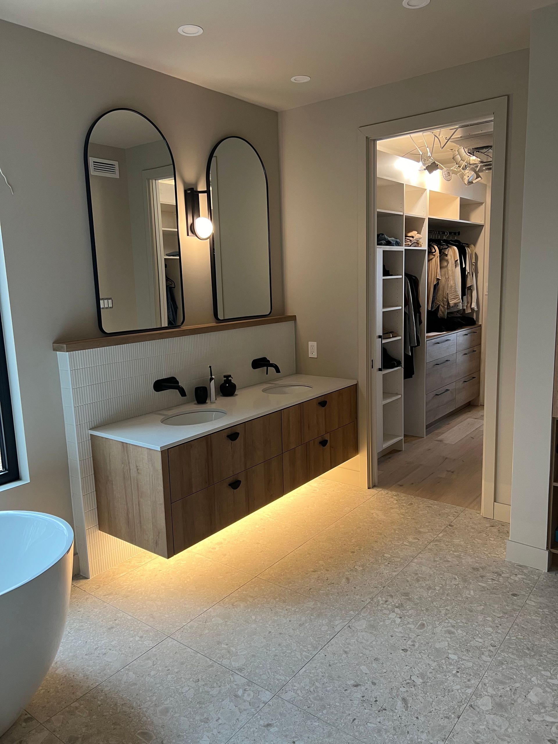 Salle de bains avec meuble-lavabo suspendu en bois, miroirs arqués et dressing visible par une porte.
