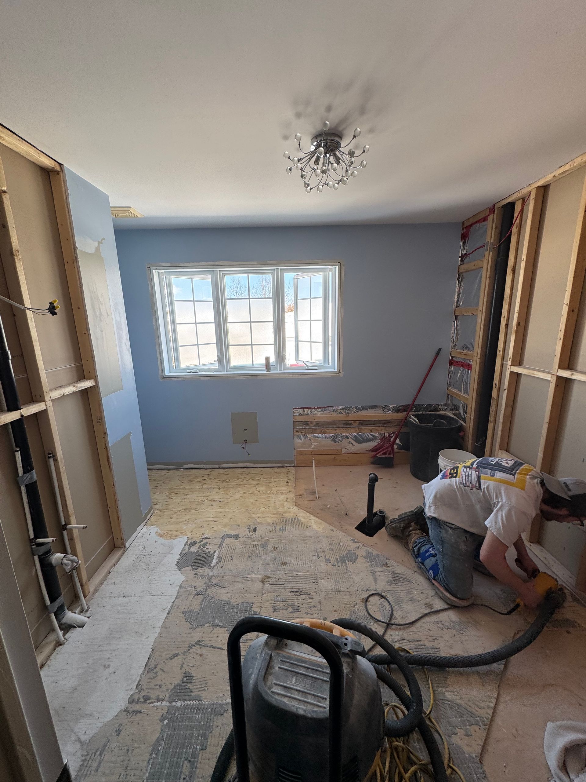 Pièce en rénovation ; murs apparents, murs bleus, fenêtre, ouvrier, aspirateur, débris au sol.