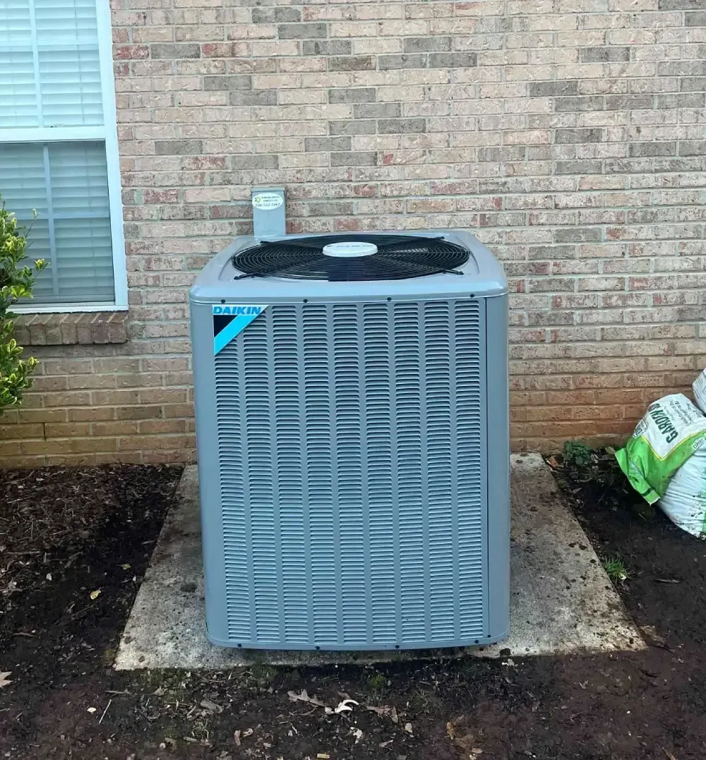 Gray outdoor HVAC unit beside a brick house wall, with a window to the left and mulch around it.