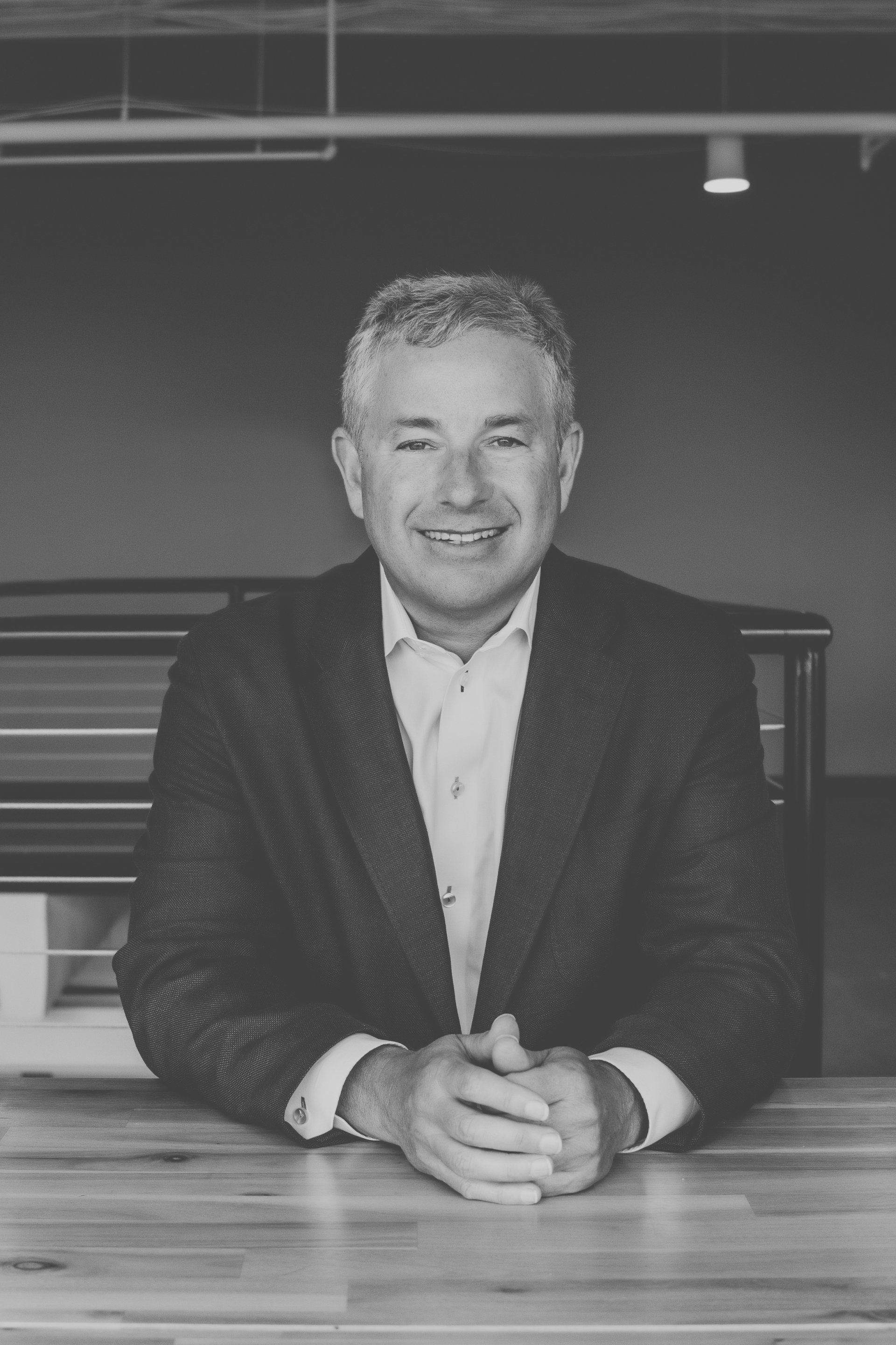 A man in a suit is sitting at a table with his hands folded.