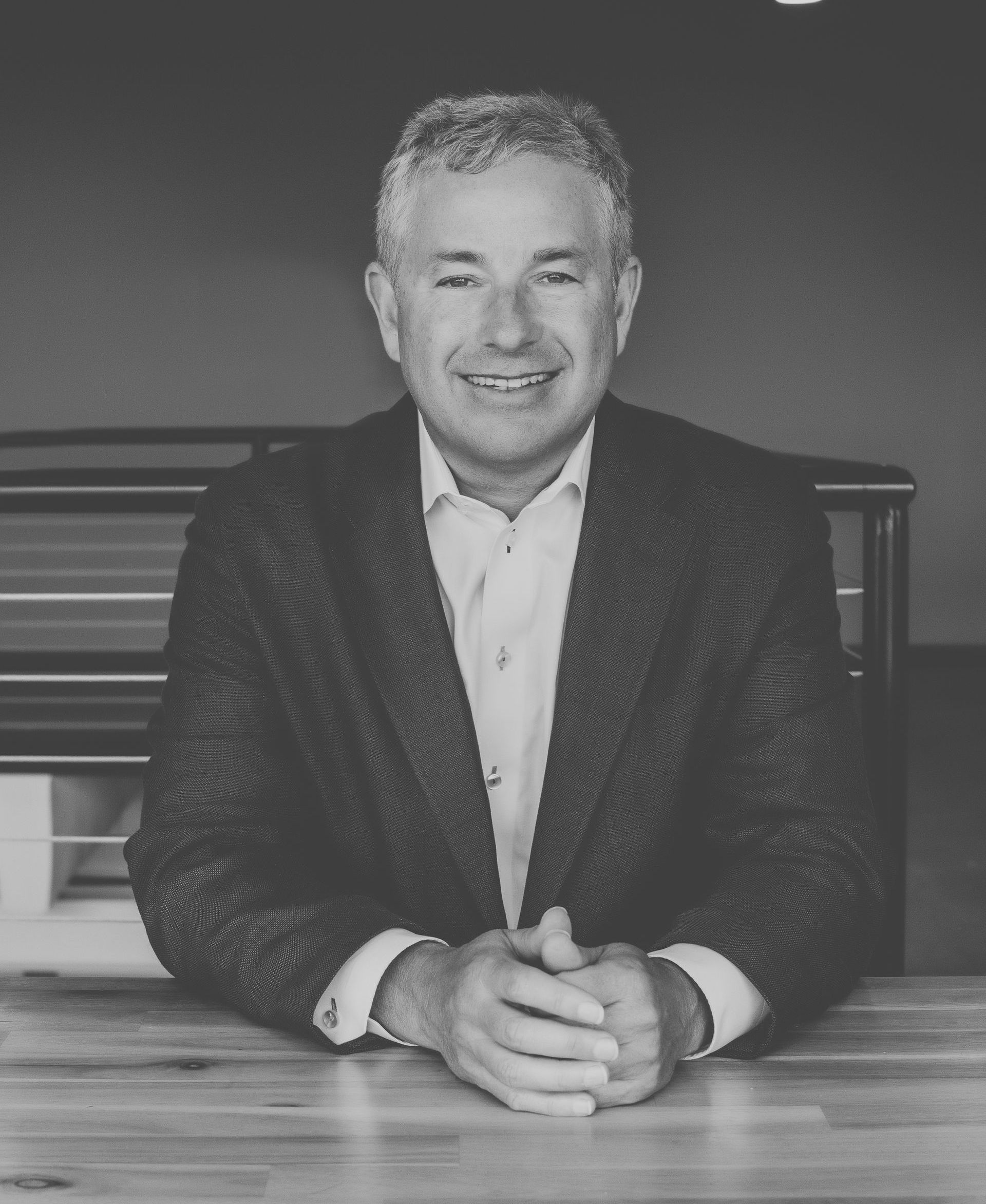 A man in a suit is sitting at a table with his hands folded.