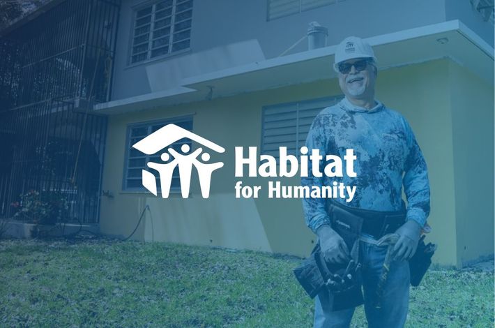 A man is standing in front of a yellow house with the habitat for humanity logo.