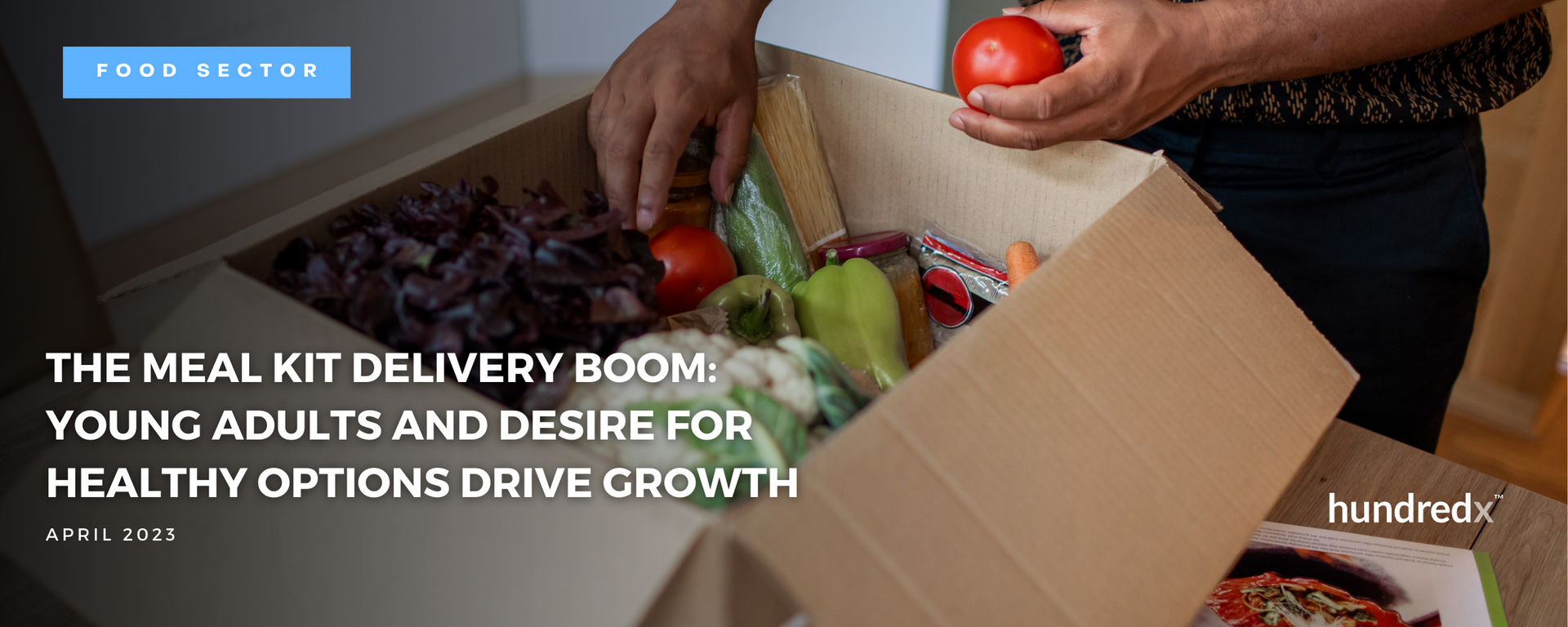 A person is holding a tomato in a cardboard box filled with vegetables.