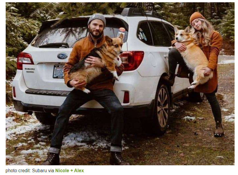 A man and a woman are holding two dogs in front of a white suv.