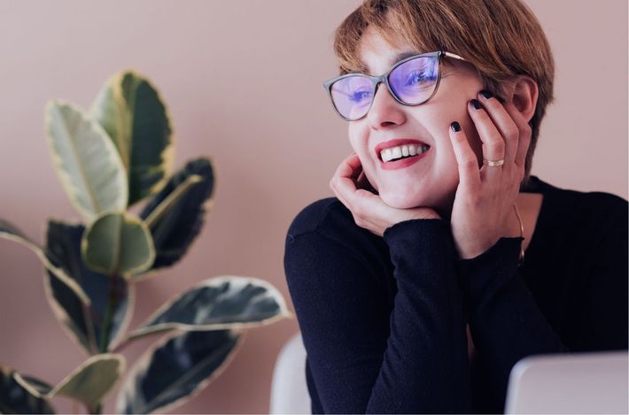 A woman wearing glasses is smiling while sitting in front of a laptop computer.