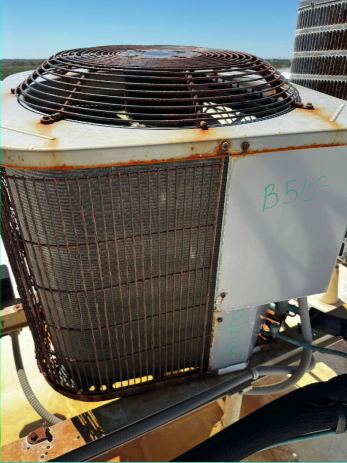 Rusty air conditioning unit on a rooftop. Metal fan cage, condenser fins, and casing show corrosion.