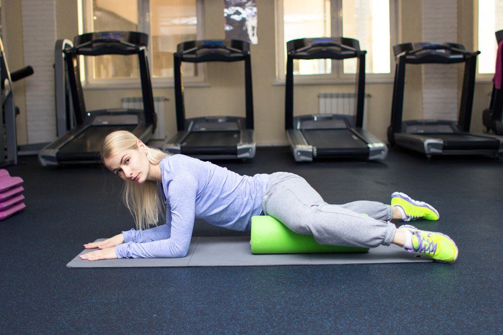 A woman is laying on a yoga mat with a green foam roller in a gym.