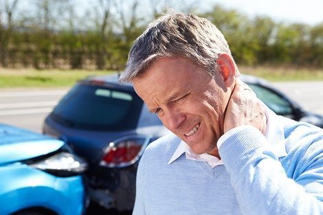 Injured man after car crash holding neck near two wrecked vehicles on roadside accident scene.