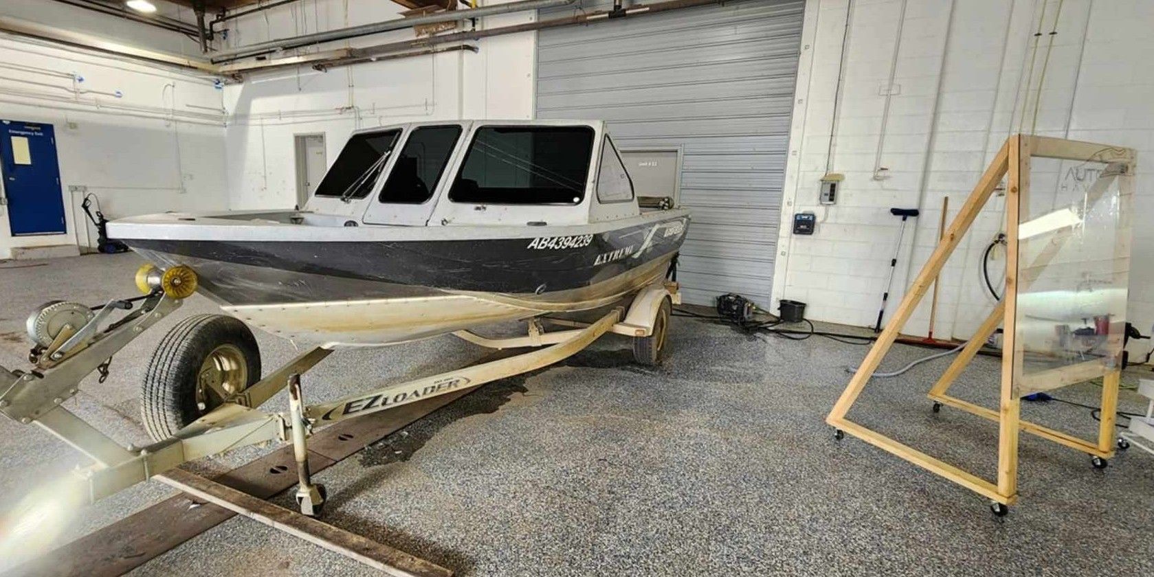 A boat on a trailer inside a building. A frame with a sheet of glass is on the right.