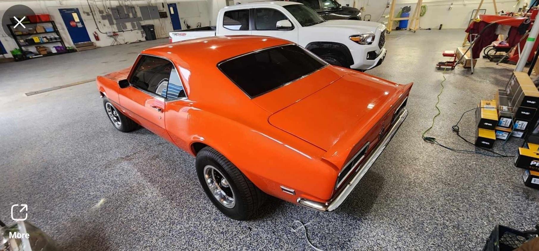 Bright orange classic car inside a garage, with a white pickup truck.