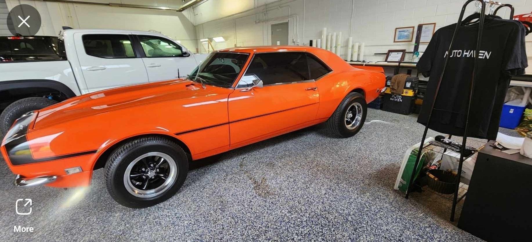 Bright orange classic car in a garage next to a white truck.  A black t-shirt hangs on a rack.
