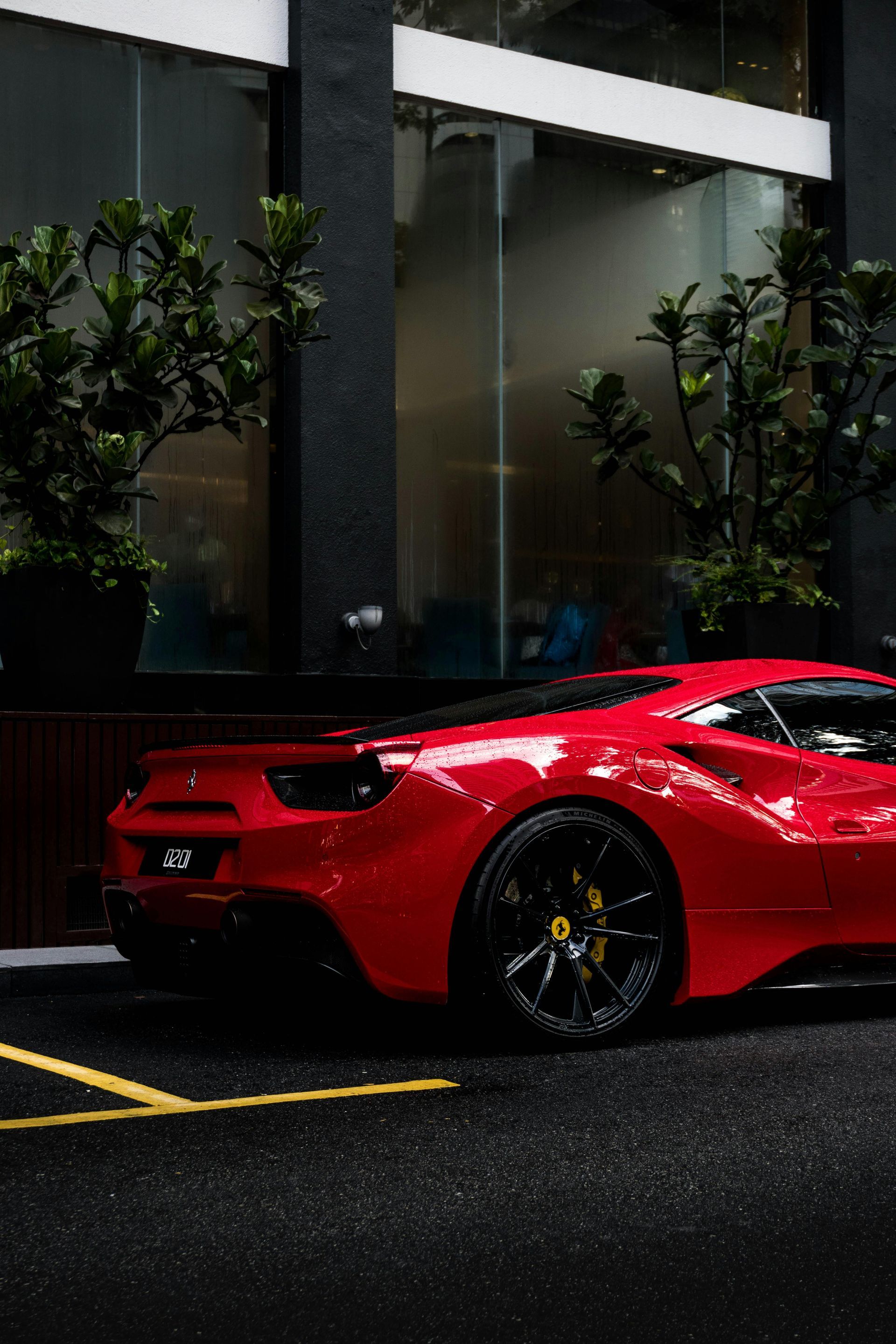 Red Ferrari sports car parked on a wet street next to a building with glass windows and foliage.