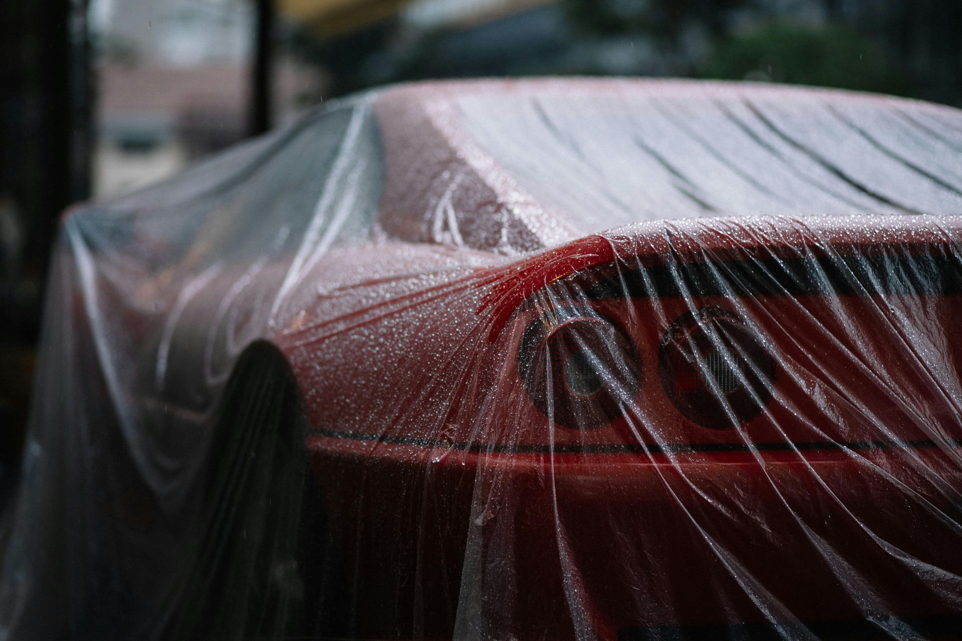 Red car covered in a wet, clear plastic sheet. Raindrops visible; setting is outdoors.