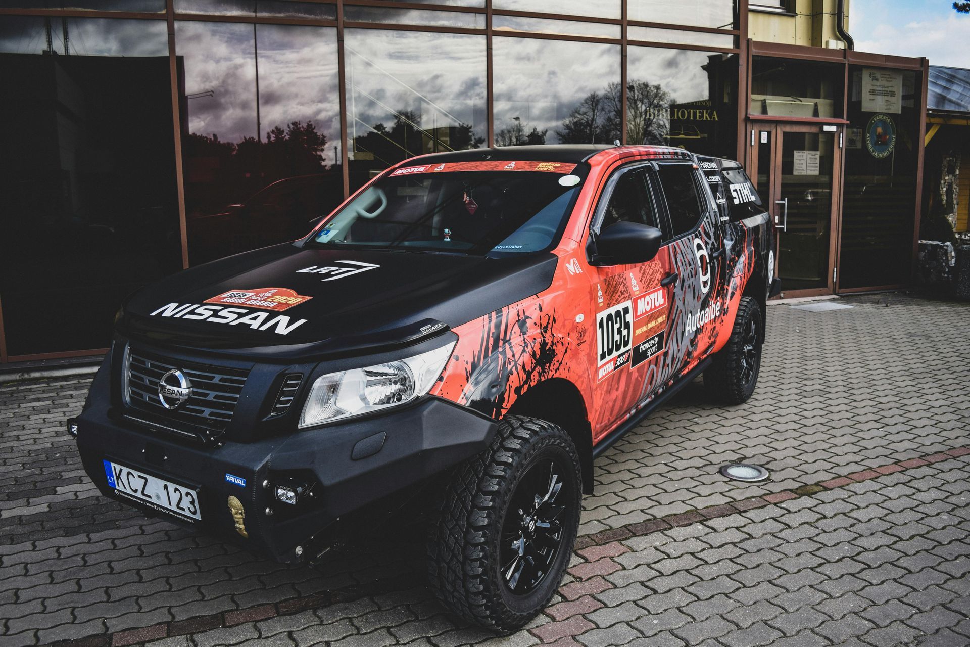 Nissan Navara pickup truck with red and black graphics, parked on pavers near a building.