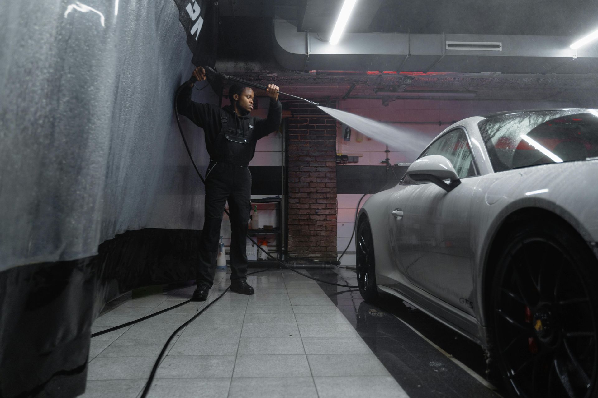 A person washes a white car with a water hose in a garage.