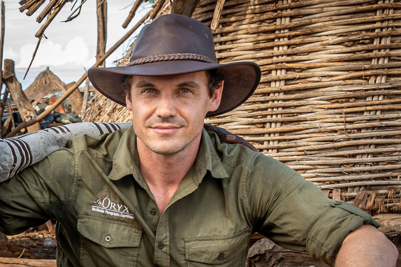 A man wearing a cowboy hat and a green shirt is standing in front of a wicker wall.