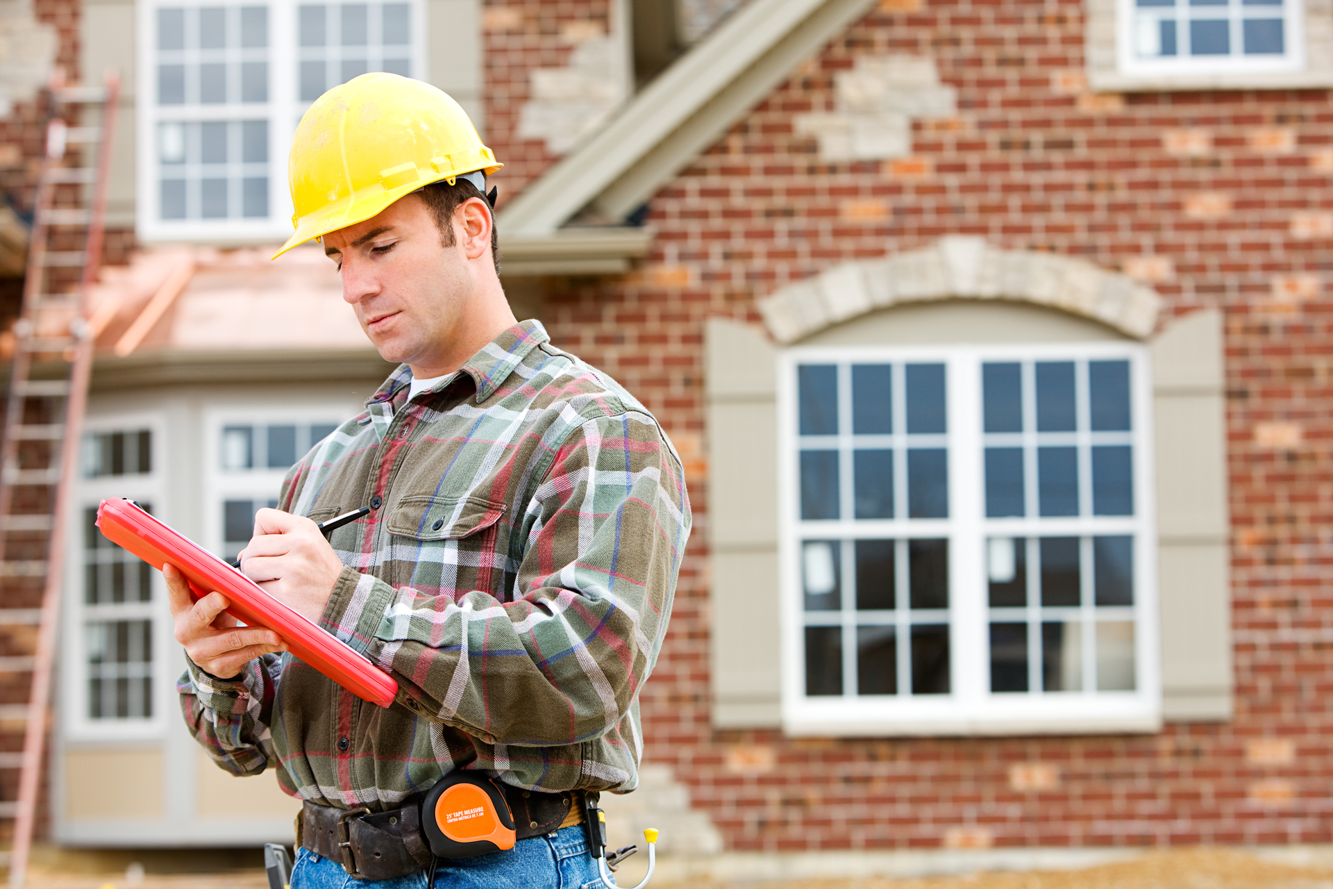 A construction worker is writing on a clipboard in front of a brick house.