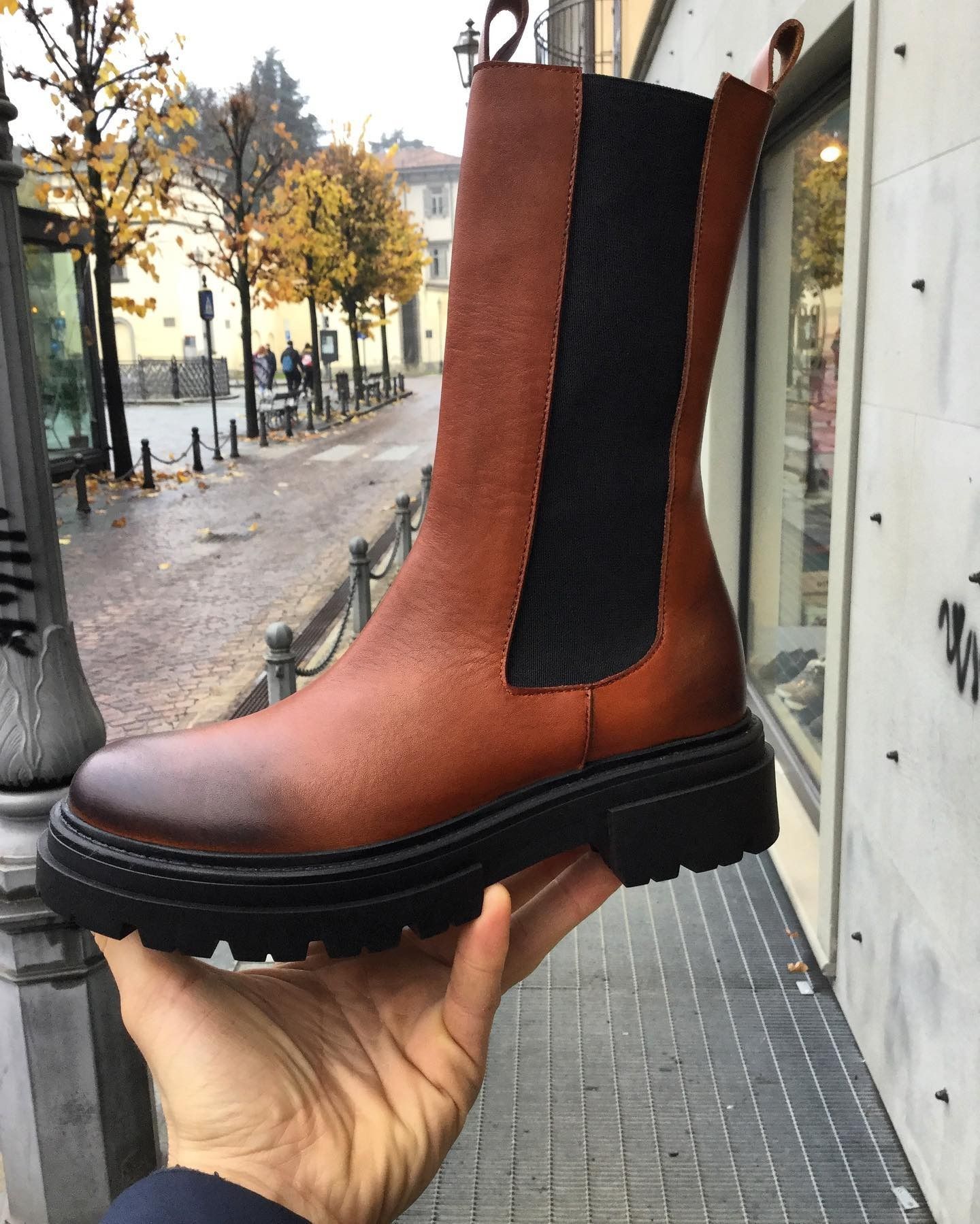 Stivale in pelle marrone con pannello laterale nero, tenuto in mano, appoggiato sul marciapiede cittadino.