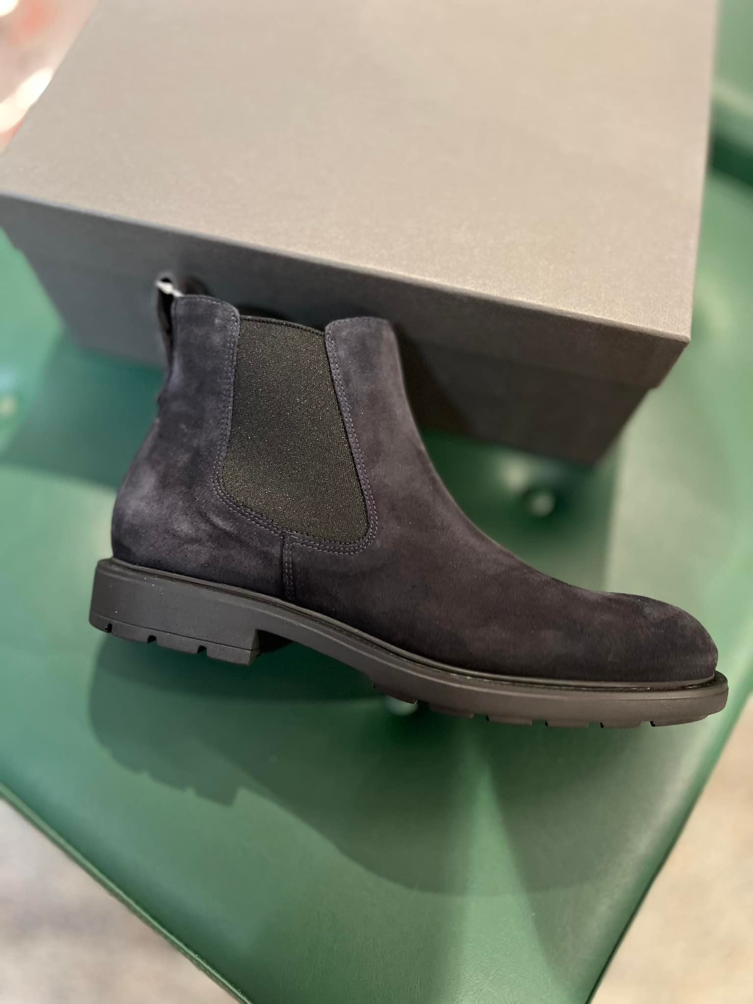 Stivaletto Chelsea in pelle scamosciata nera su una superficie verde, con una scatola grigia dietro.