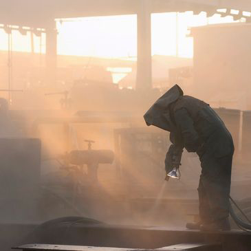 A man is sandblasting a piece of metal in a factory.